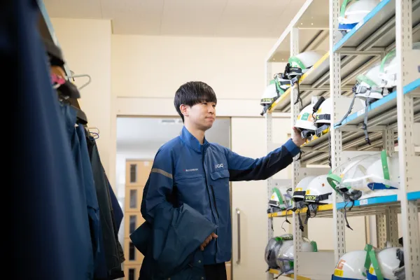 作業服を着た若い男性社員が、ヘルメットや防護具が並ぶ棚から装備を選んでいる社内倉庫の様子