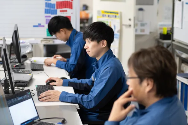 作業服を着た社員がオフィス内のデスクでパソコン操作を行い、周囲で同僚が資料確認や作業を進めている業務風景