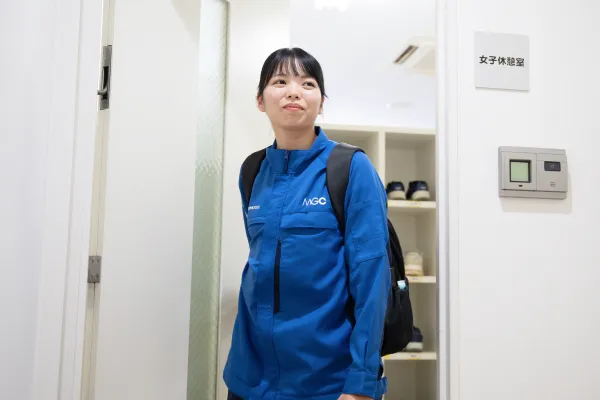 作業服を着た女性社員が更衣室の入口に立ち、リュックを背負って出勤・準備をしている社内施設の様子