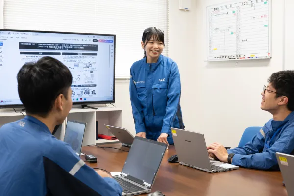 会議室で女性社員が立って説明し、周囲の同僚がノートパソコンを開いて話を聞く打ち合わせ風景