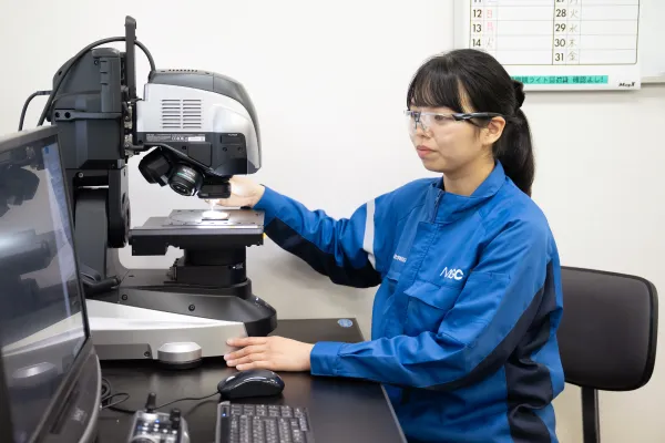 保護メガネを着用した女性社員が顕微鏡や測定装置を操作し、デスク上で分析作業を行っている研究・検査の場面