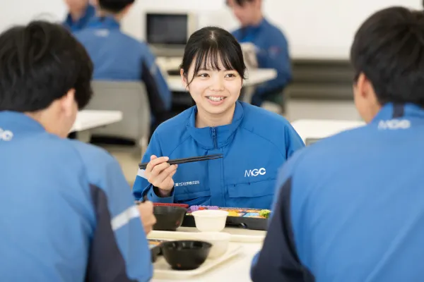 社員食堂で作業服を着た女性社員が同僚と向かい合い、箸を持って会話を楽しみながら昼食を取っている様子